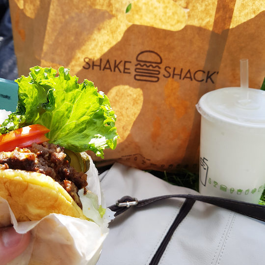 shake shack burger being held in the hand with shake shack brown bag and soda in the background