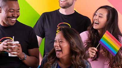 four people smiling and looking at each other. Two are holding pride flags, one is holding a shake. There is a rainbow wall behind them.