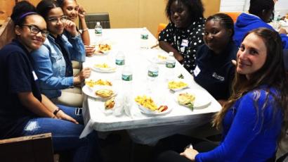 six women sitting around a table with food on it, smiling at the camera. 