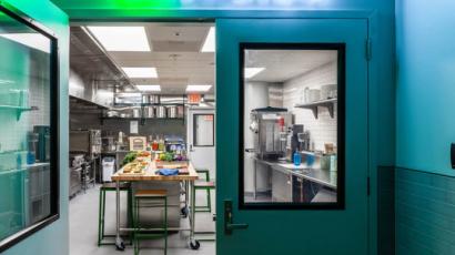Picture of two swinging doors. One is open, allowing us to see an industrial kitchen. The words Innovation Kitchen are spelled out above the door in neon next to a shake shack logo.