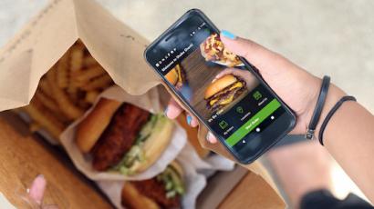 hand holding a phone with the shake shack app on it. Below it is a bag with fries and burgers in it.