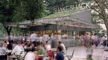 Shake Shack storefront with outdoor seating in front of it. There is a line at the window and people sitting at every table.