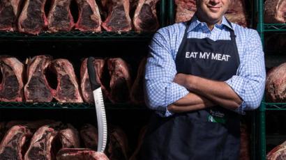 Pat LaFrieda in an apron, with arms crossed, standing in front of racks full of meat