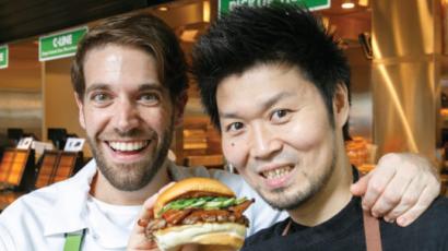 Two people in aprons smiling at the camera. One is holding a burger.
