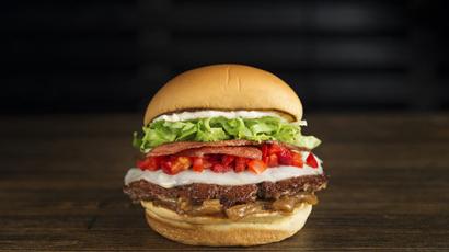 burger held in two hands. Behind it on the table is another burger and fries.