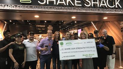 Group of smiling people, some of them shake shack employees, holding a check for $632,544 made out to Share Our Strength. They are standing in from of a Shake Shack storefront.