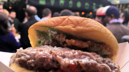 close up of a burger with a shake shack storefront behind it.