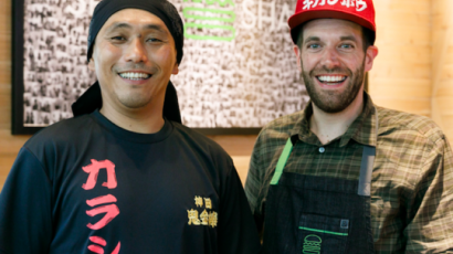 two people smiling at the camera with a burger on the table in front of them. One person is wearing an apron.