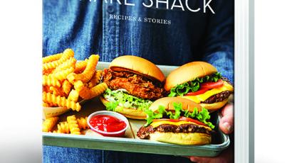 Hard covered book titled Shake Shack. There is a picture of a someone holding a tray with three burgers and fries on it.